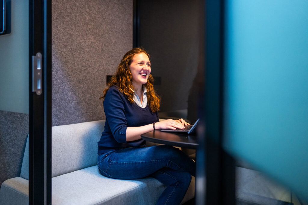 Person smiling while working inside a meeting pod on their laptop.