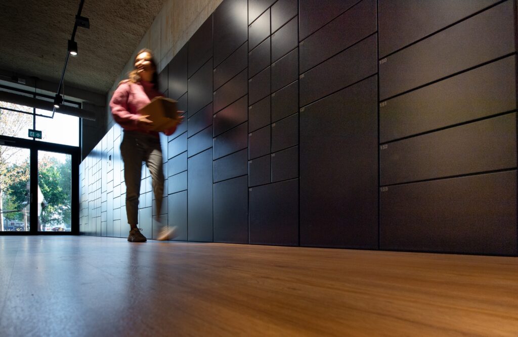 Person carrying a parcel next to a bank of smart lockers in a corporate environment.